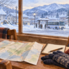地図と防寒用品を広げた机から雪の町のバスを見る旅行準備のイラスト